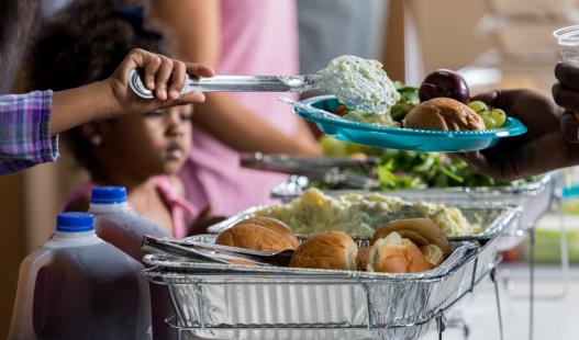 A volunteer ladles healthy food on a plate in a soup kitchen while a young African-American girl looks on.