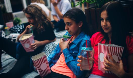 A group of children enjoy popcorn and lemonade as they watch a movie on blankets in a backyard.