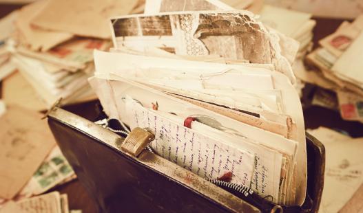 Old handwritten letters and papers are stuffed inside of a vintage handbag on a desk strewn with letters.