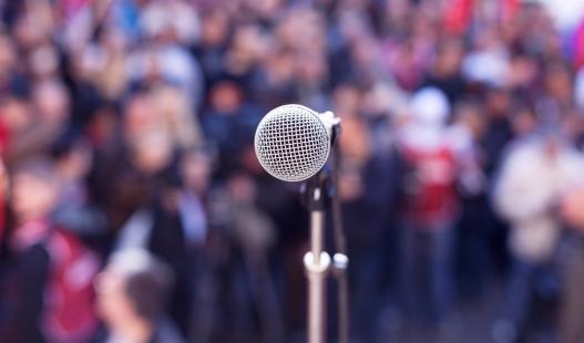 Close-up of a microphone as seen from a stage, with a blurred background of spectators at an outdoor rally