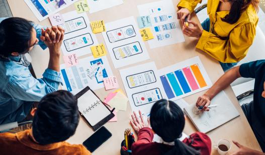 Birds eye view of a group of people sitting at a table, looking at paper prototypes and other mobile app documentation