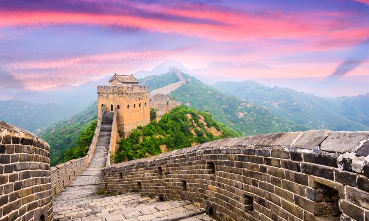 View of China's Great Wall, with a pink sky and mountains behind it.